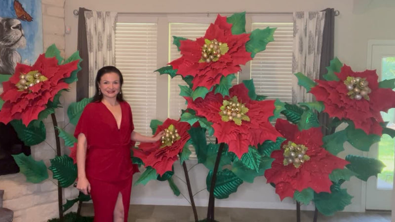 Woman in a red dress standing next to large paper poinsettias in a room with a window and curtains.